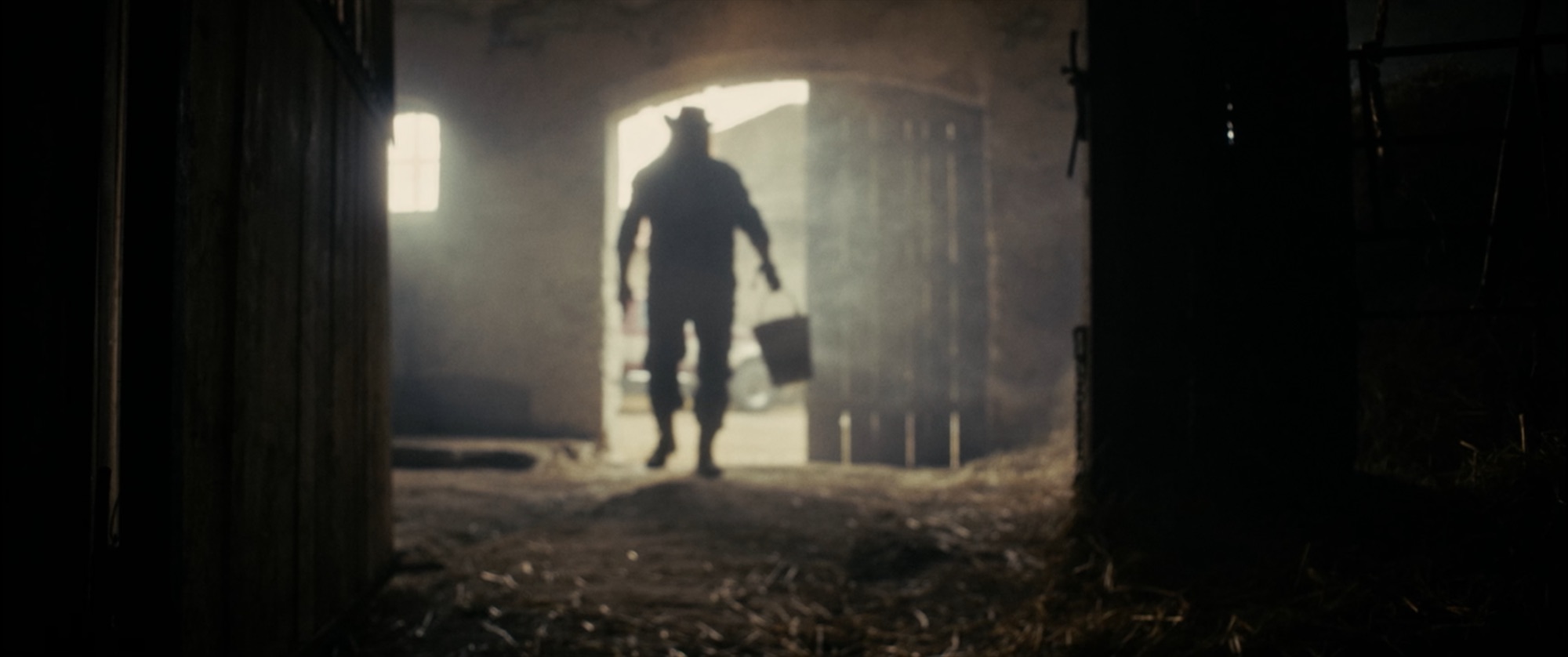 Silhouette of a figure in a cowboy hat carrying a bucket through a smoky, backlit barn doorway - Amon Amarth